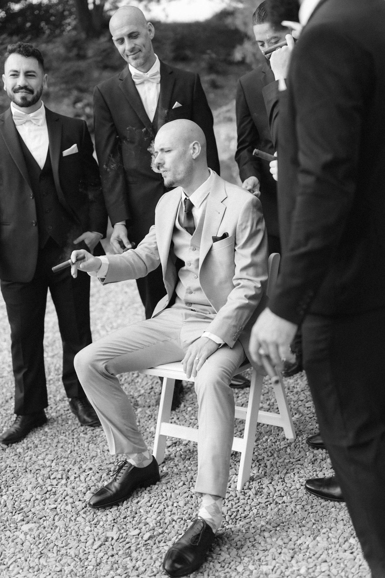 A groom sitting on a chair looking down thoughtfully while holding a cigar, surrounded by a group of formally dressed men.