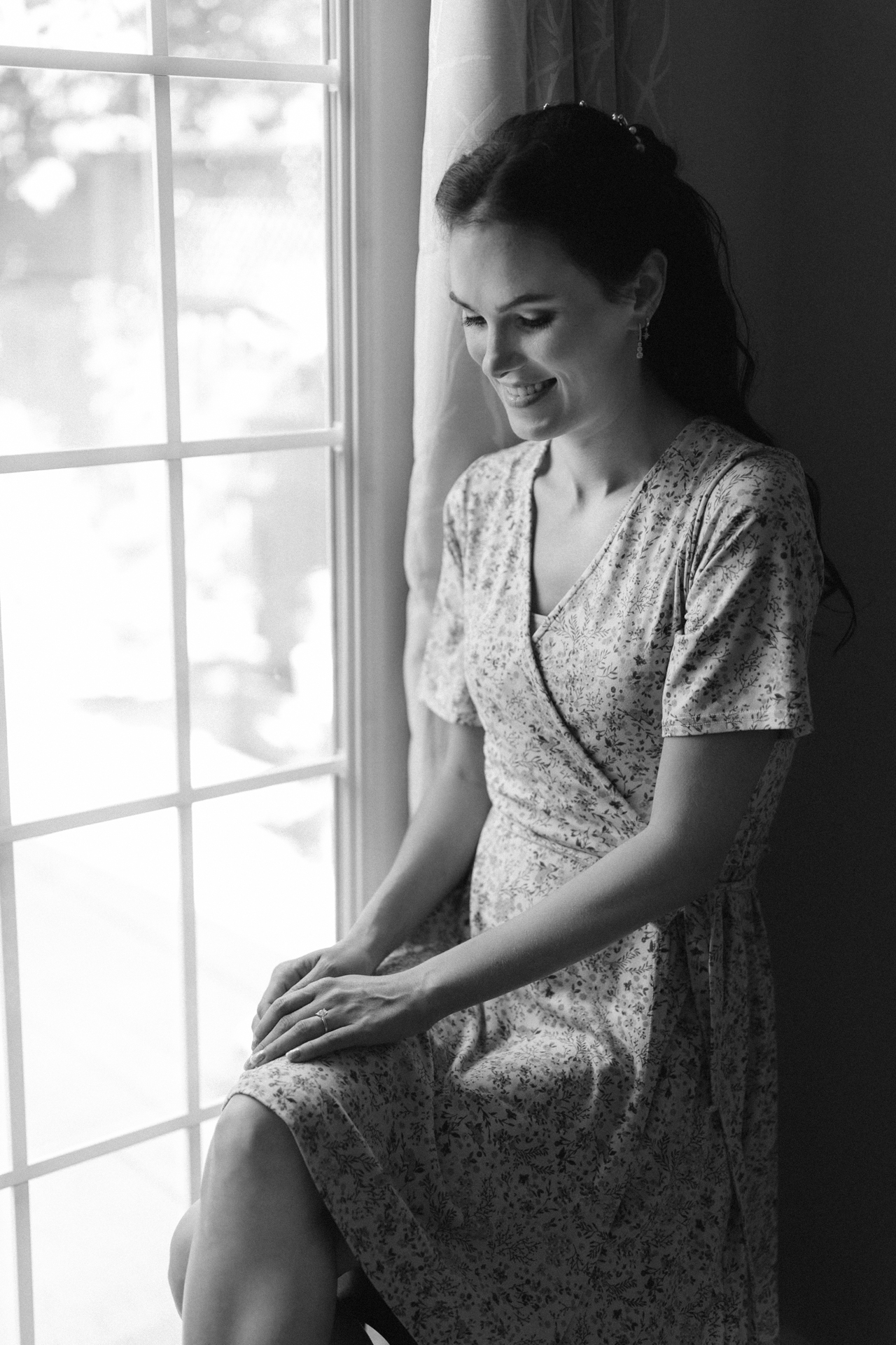 Bride in a floral dress smiling softly while sitting beside a window with sheer curtains.