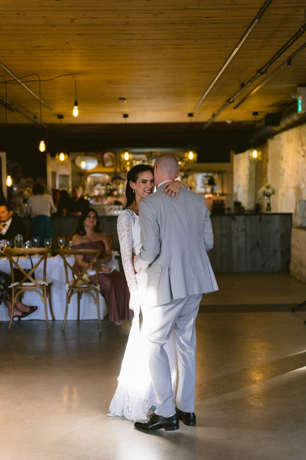 A couple sharing a dance in a warmly lit venue, with guests seated in the background