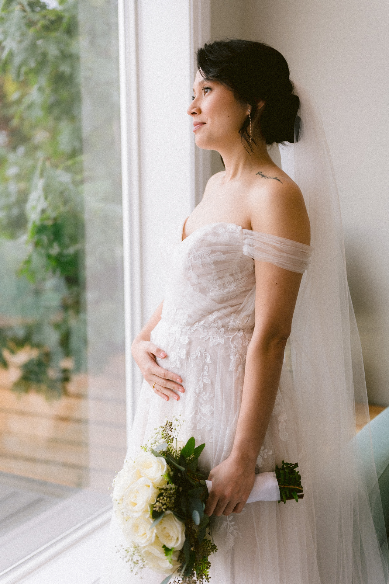 A bride in an off-the-shoulder lace dress with a veil, holding a bouquet, gazes out a window