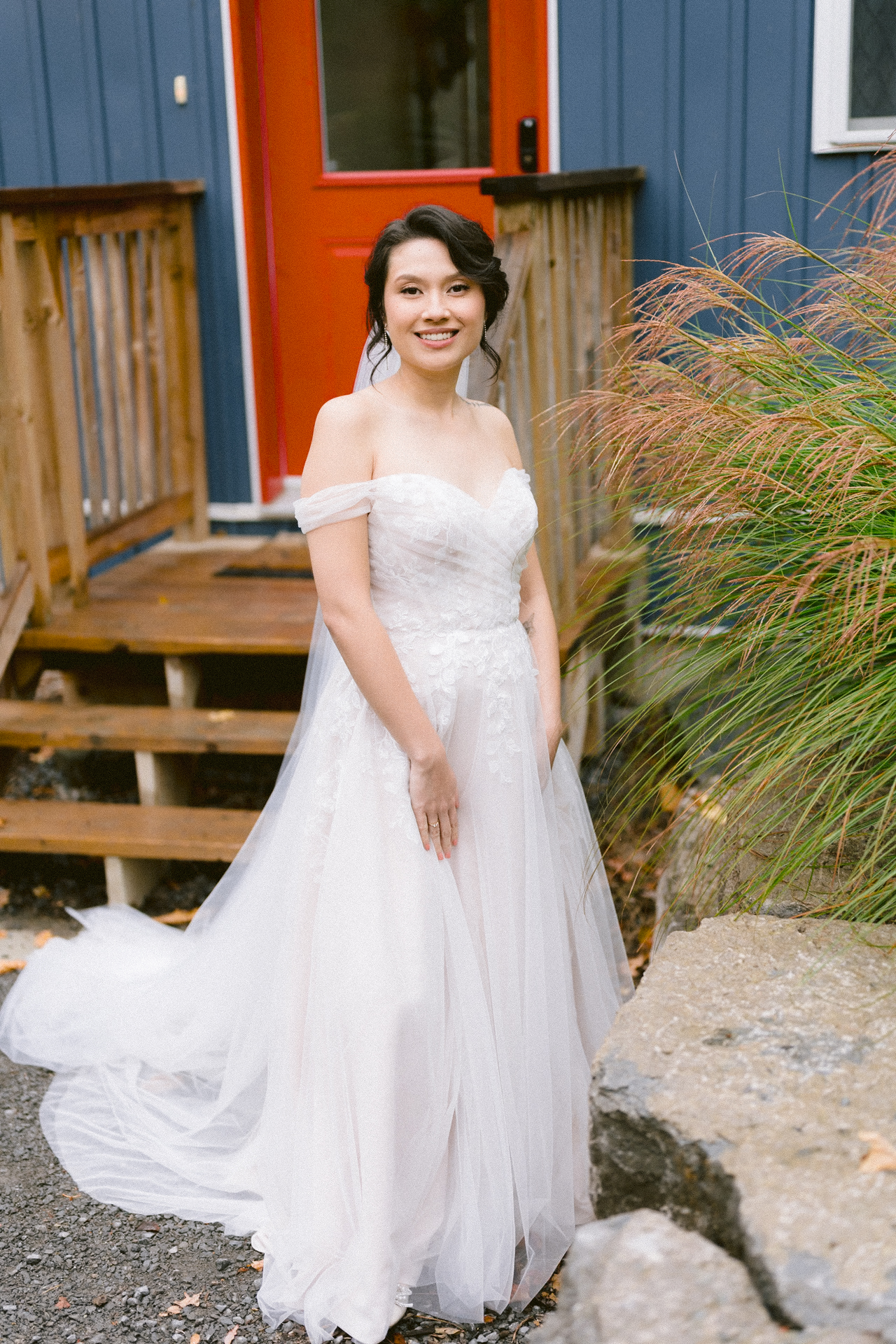 A bride in a white gown in front of a blue house with a red door.