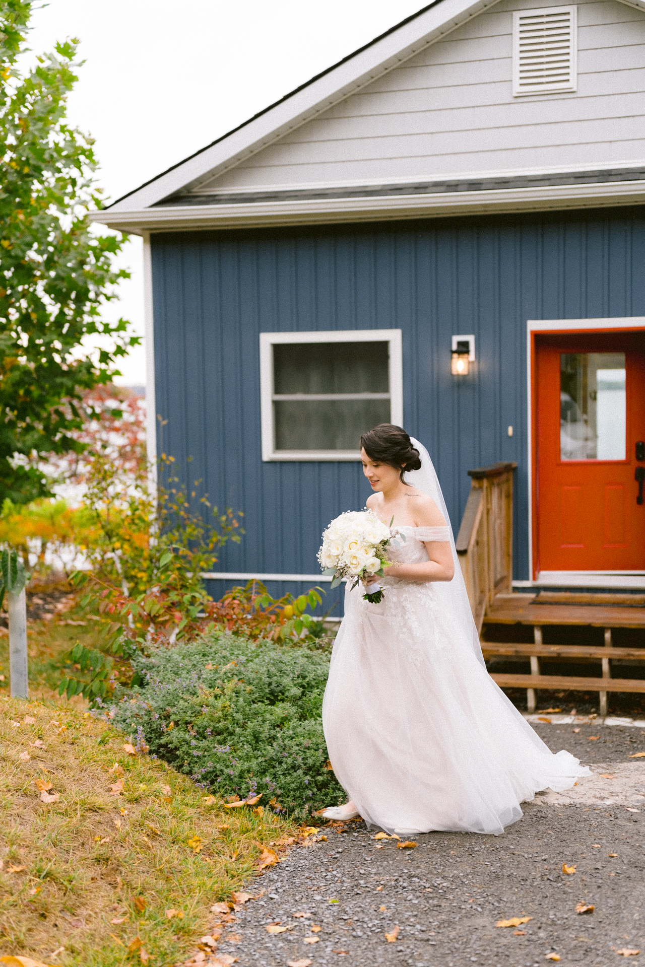 A bride in a white gown holding a floral bouquet stands in front of a blue house with a red door.