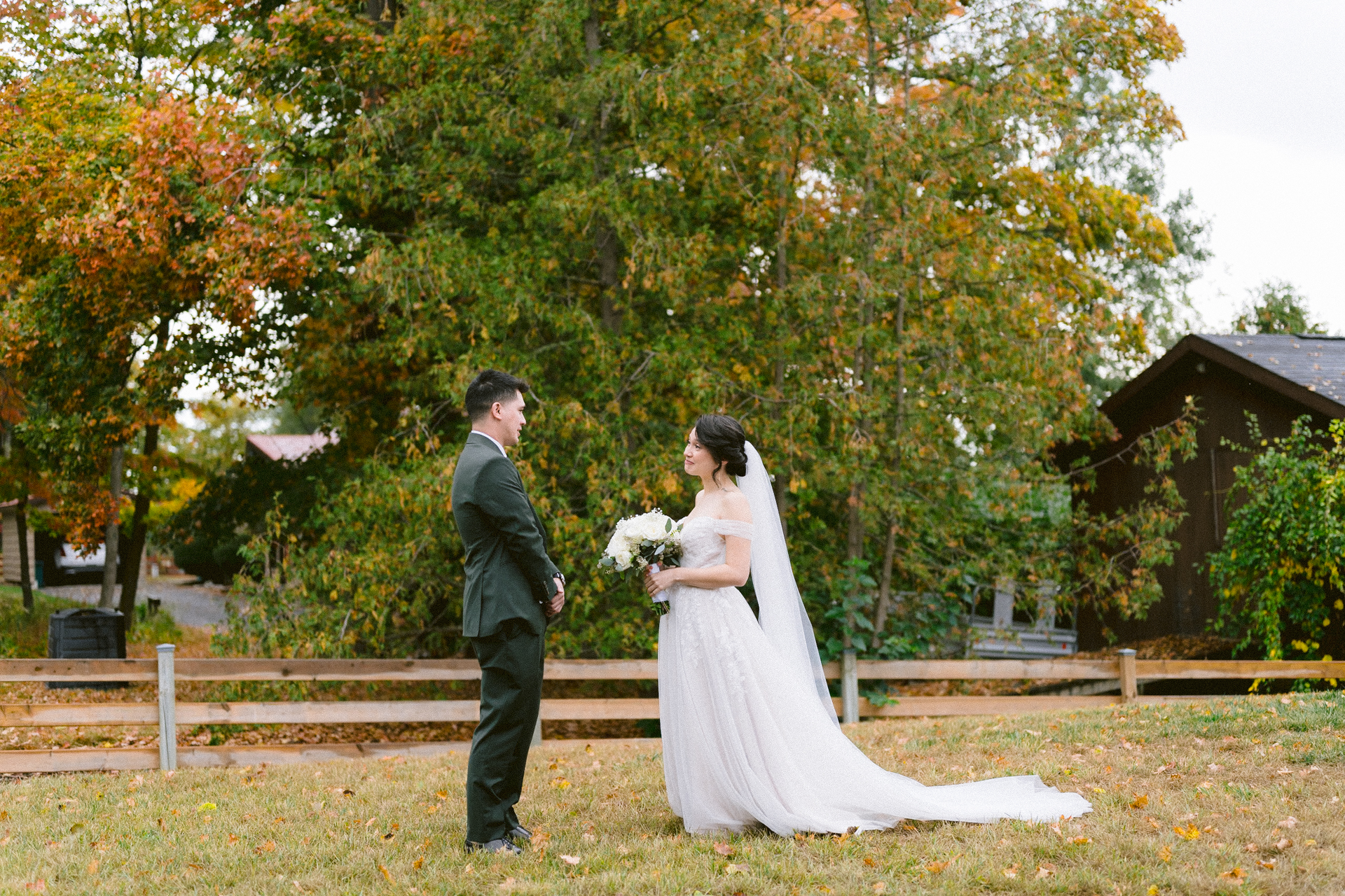 A bride and groom standing together on a lawn with autumn foliage in the background.