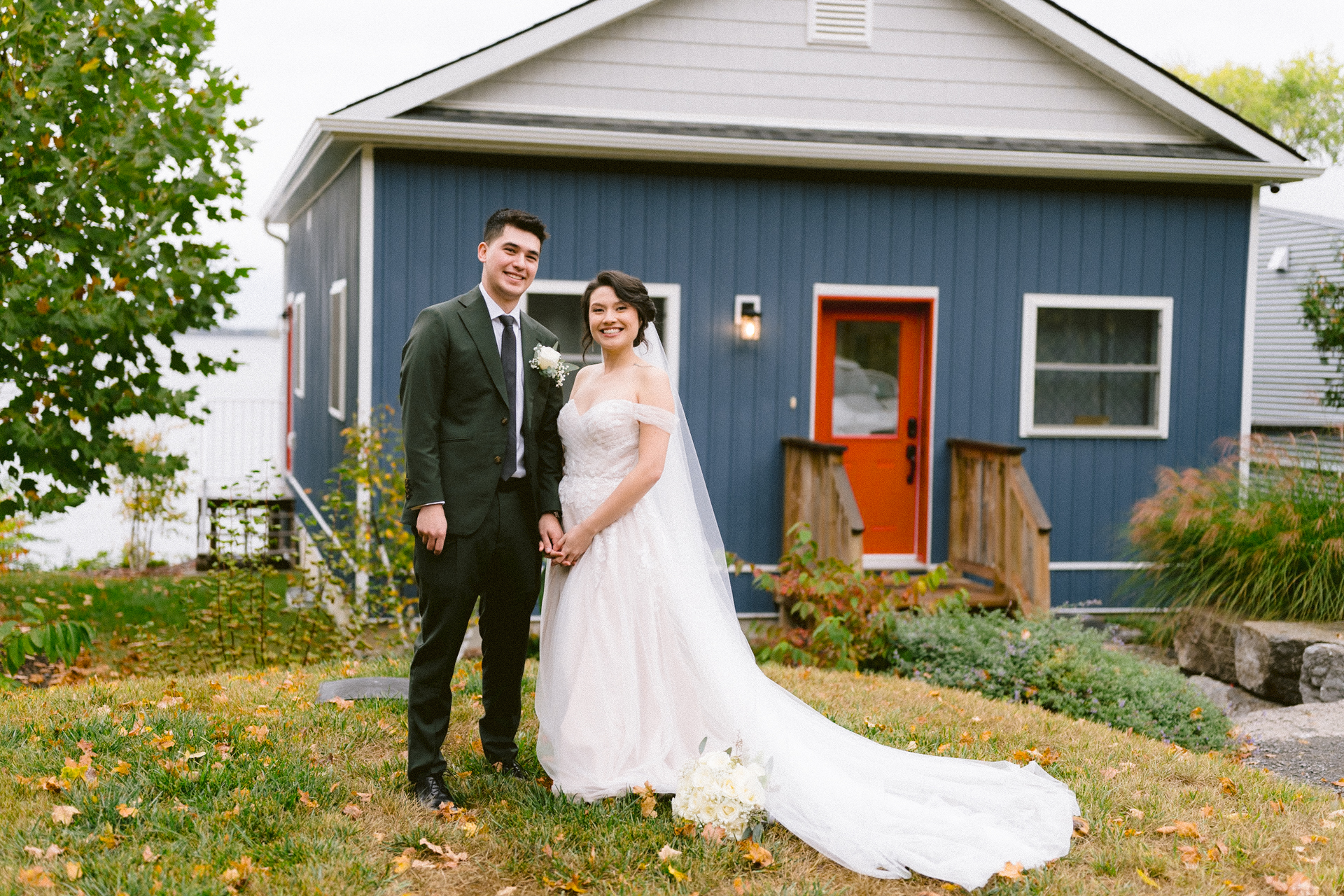 A smiling couple in wedding attire posing in front of a blue house with a red door