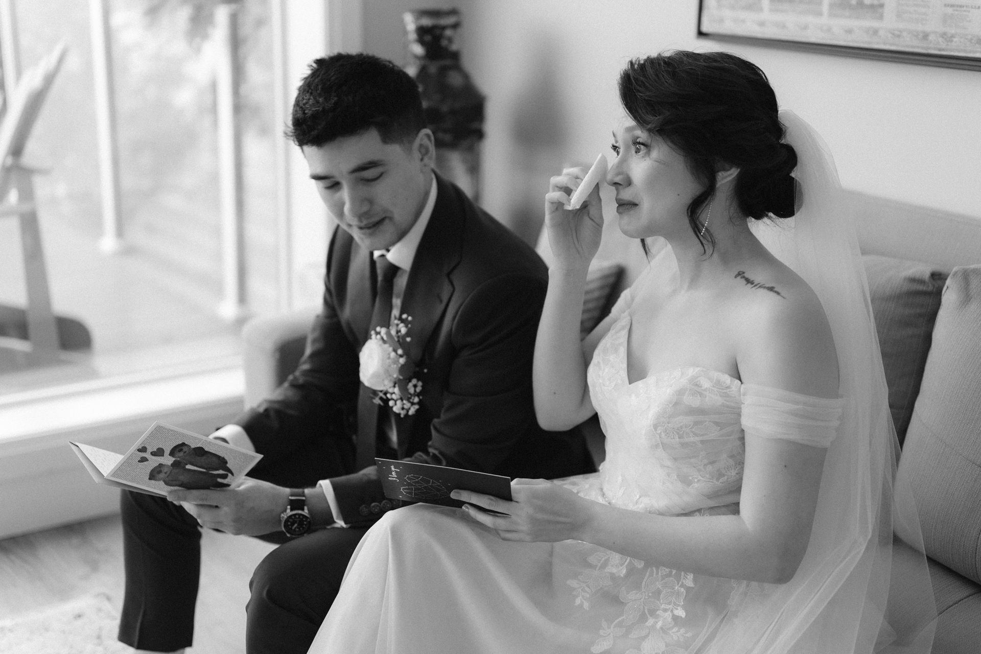 A bride and groom sitting on a sofa, reading heartfelt notes, with the groom appearing to wipe a tear and the bride smiling