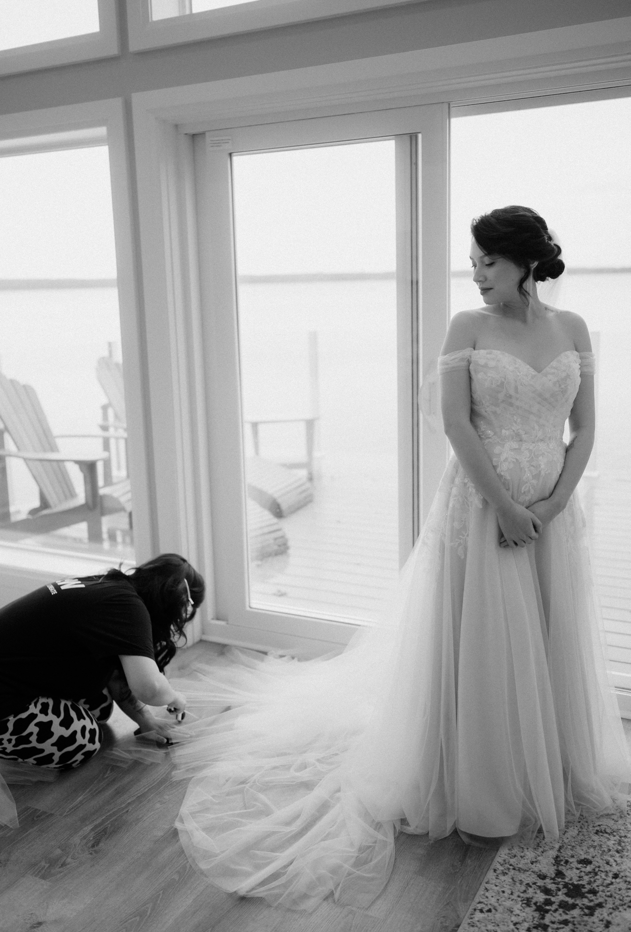 A bride stands near a window overlooking a lake while another person adjusts the train of her dress.