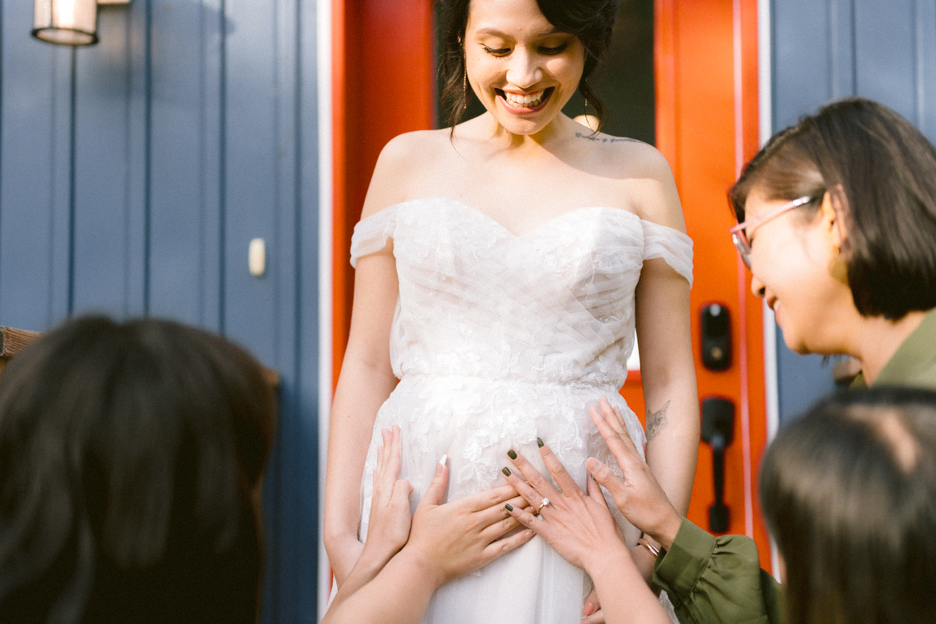 A smiling bride in a white dress is being greeted by guests touching her pregnant belly