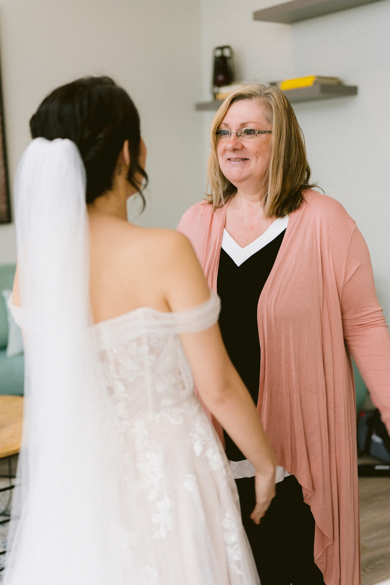 A bride in her wedding dress facing a smiling woman, likely a relative or friend, in a living room setting