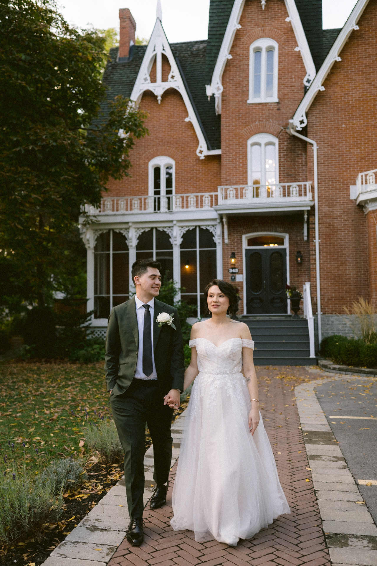 A couple in wedding attire holding hands in front of a victorian-style house