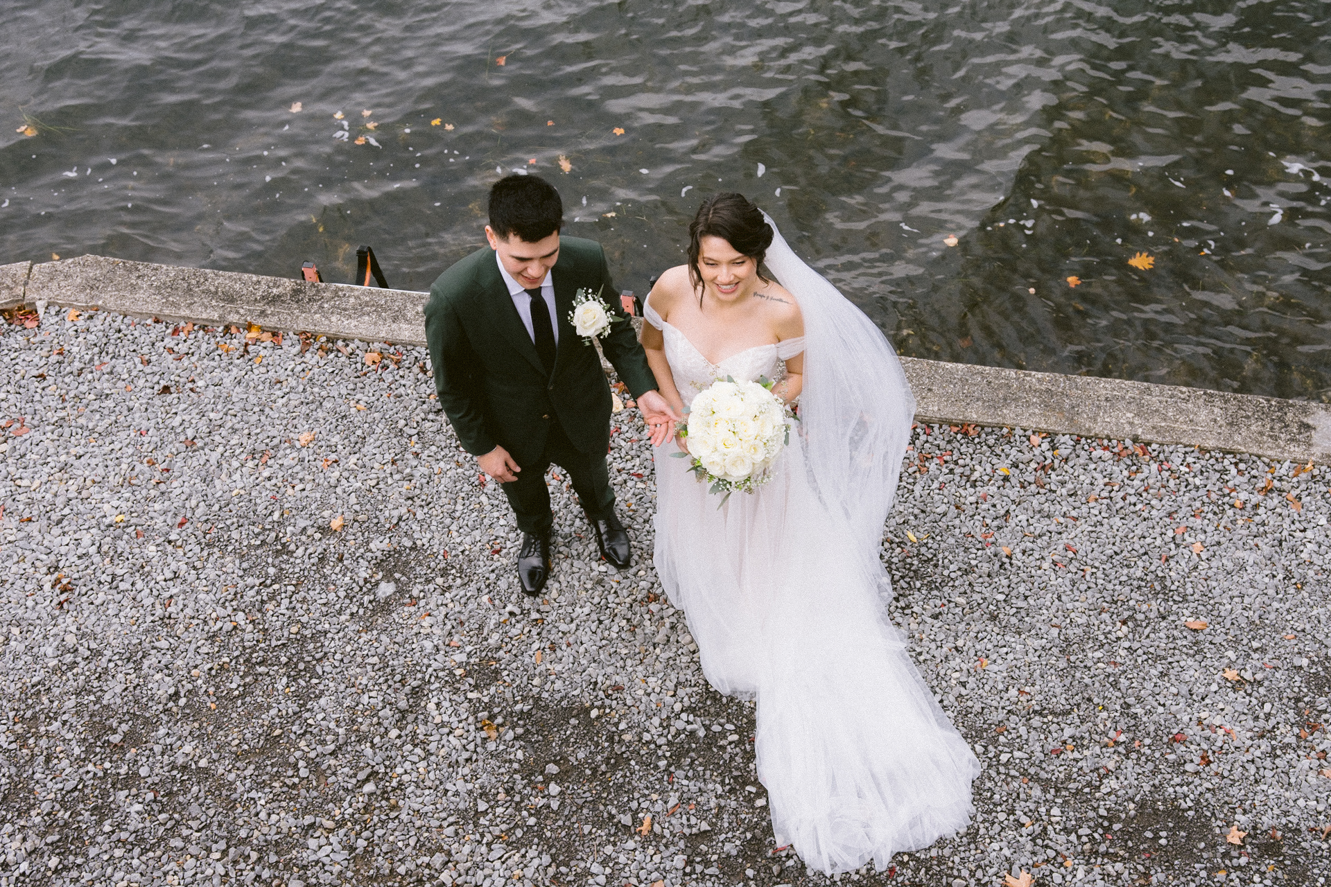 A just-married couple walking hand-in-hand along a pebbled lakeshore.