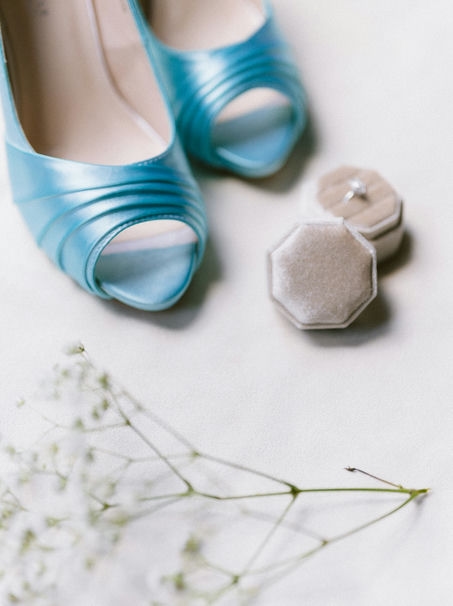 A pair of blue bridal shoes positioned next to a small ring box and delicate white flowers on a light background