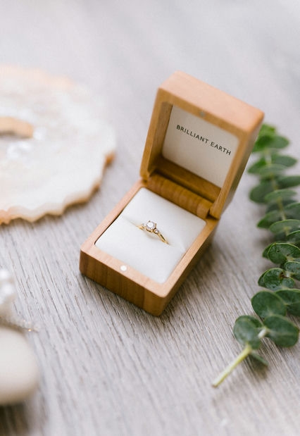 Gold engagement ring presented in a wooden box on a textured surface with greenery in the background, capturing the essence of a fall wedding