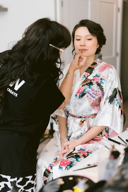 A makeup artist applying lipstick to a woman seated in a robe with a floral pattern for her fall wedding