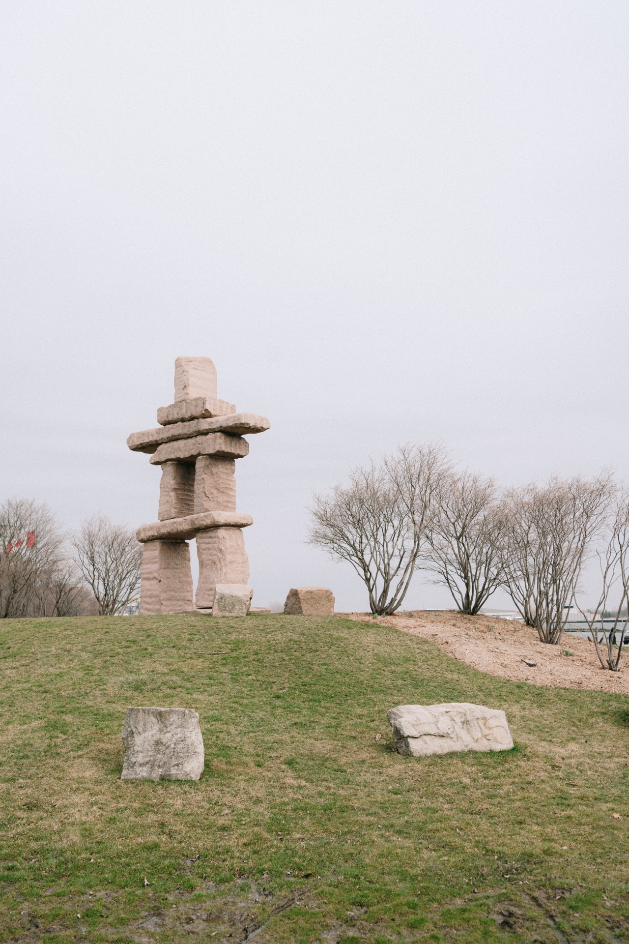 A stone inukshuk structure standing in a grassy area of Trillium Park with leafless trees and an overcast sky in the background