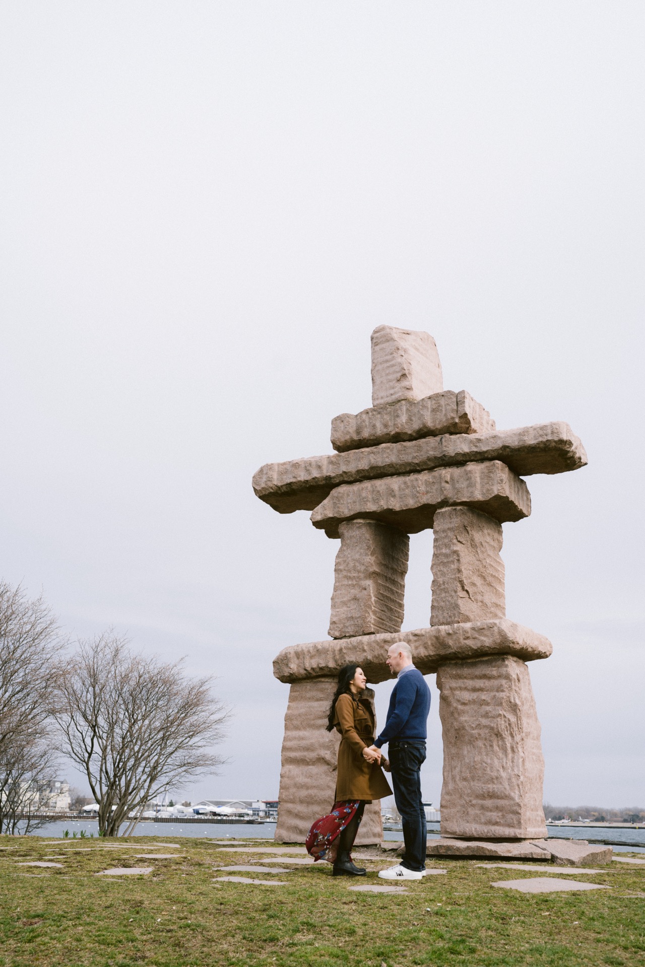 A couple stands holding hands during their engagement session in front of a large inukshuk structure at Trillium Park