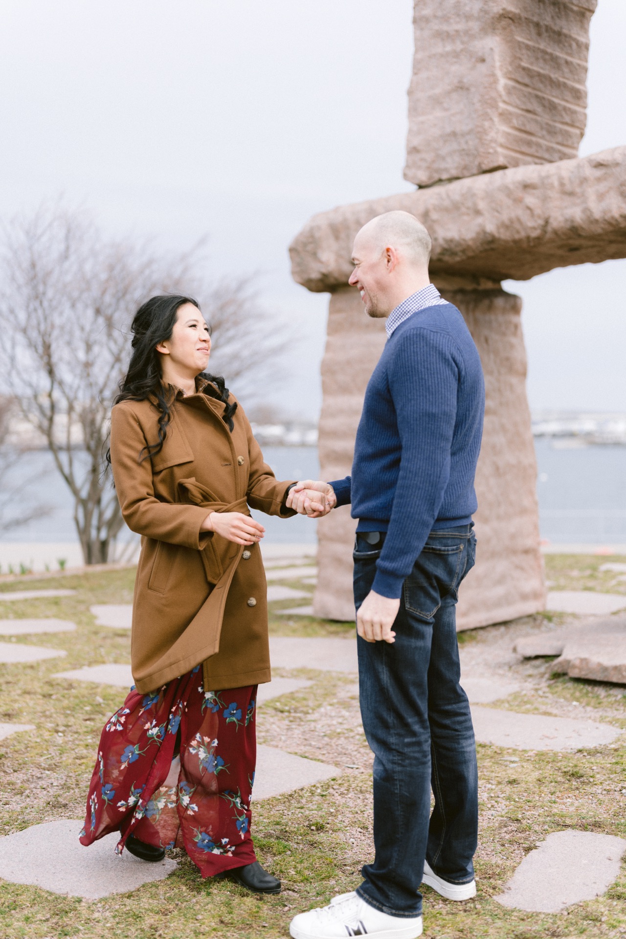 A couple holding hands standing face-to-face outdoors near a stone structure.