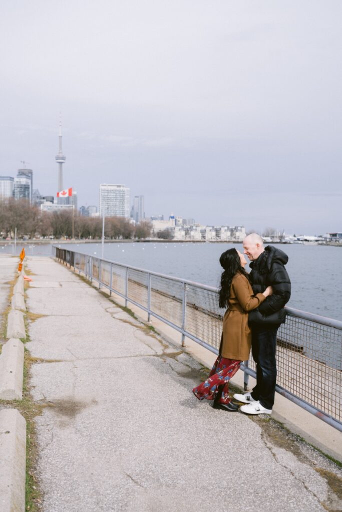 A couple sharing a romantic embrace by the waterfront with a city skyline in the background.