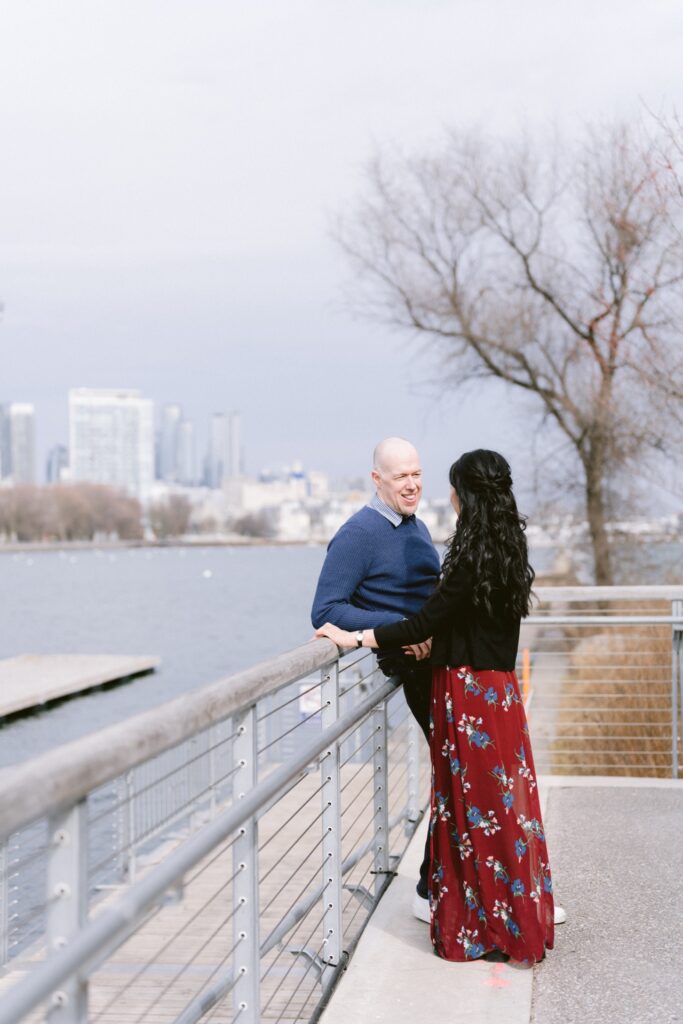 A couple smiling at each other on a waterfront promenade with a city skyline in the background.