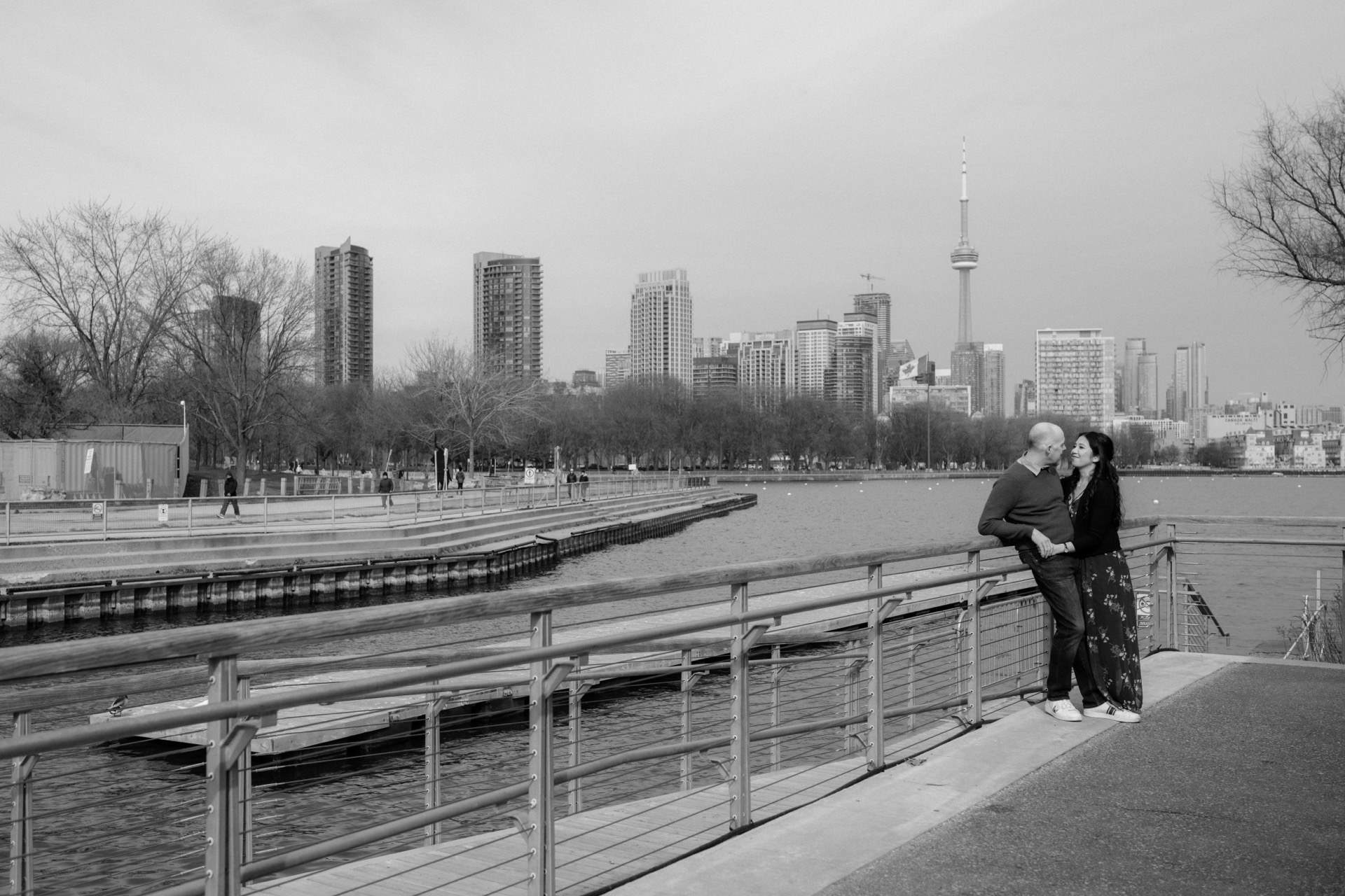 A couple embraces near a waterfront with a city skyline in the background.