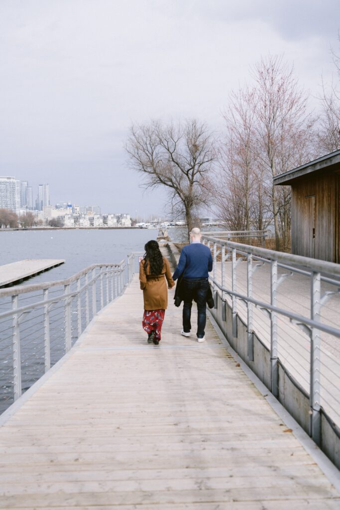 Two people walking on a wooden boardwalk beside a lake with a city skyline in the distance.