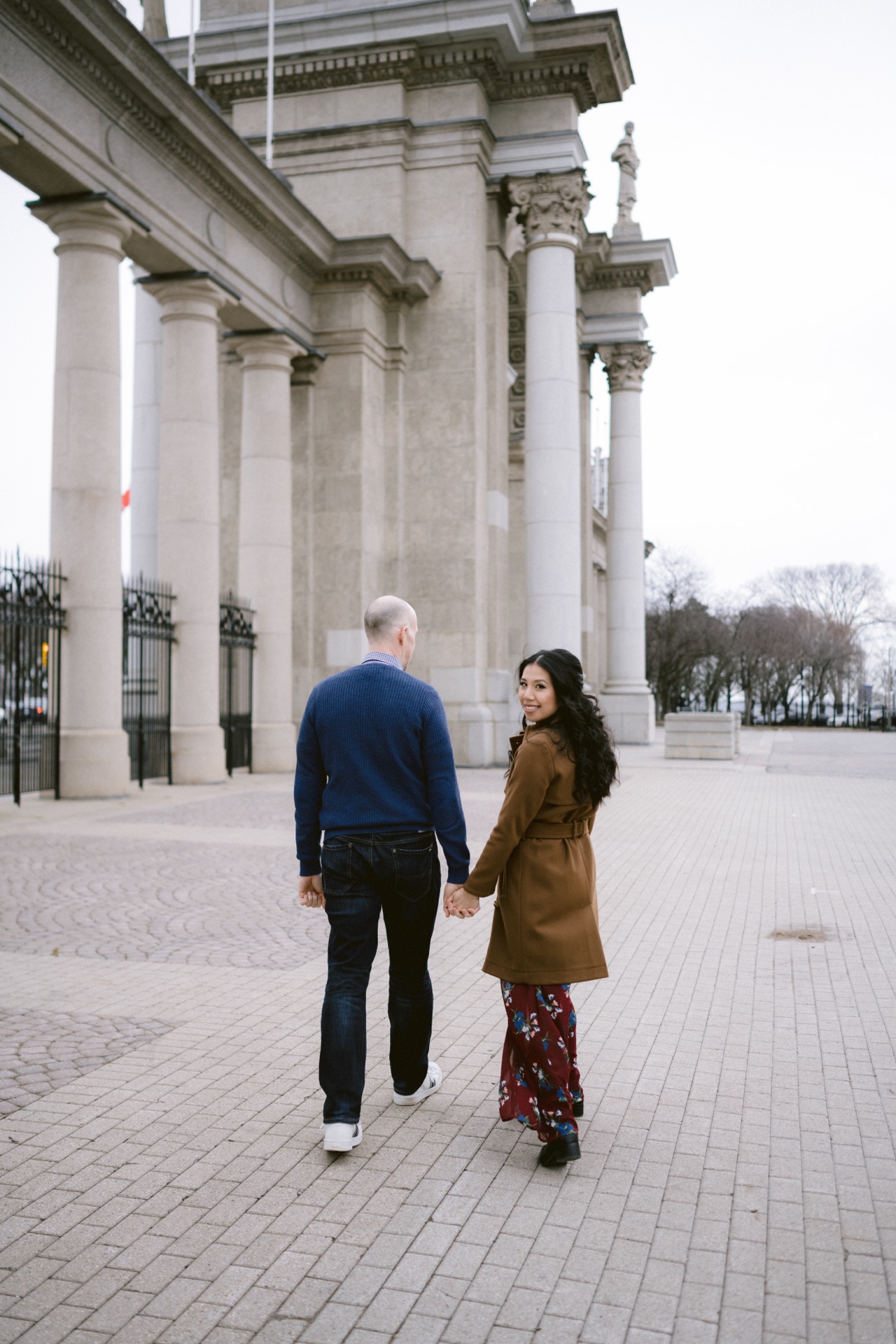 A couple holding hands while walking away on a paved area near a large building with columns.