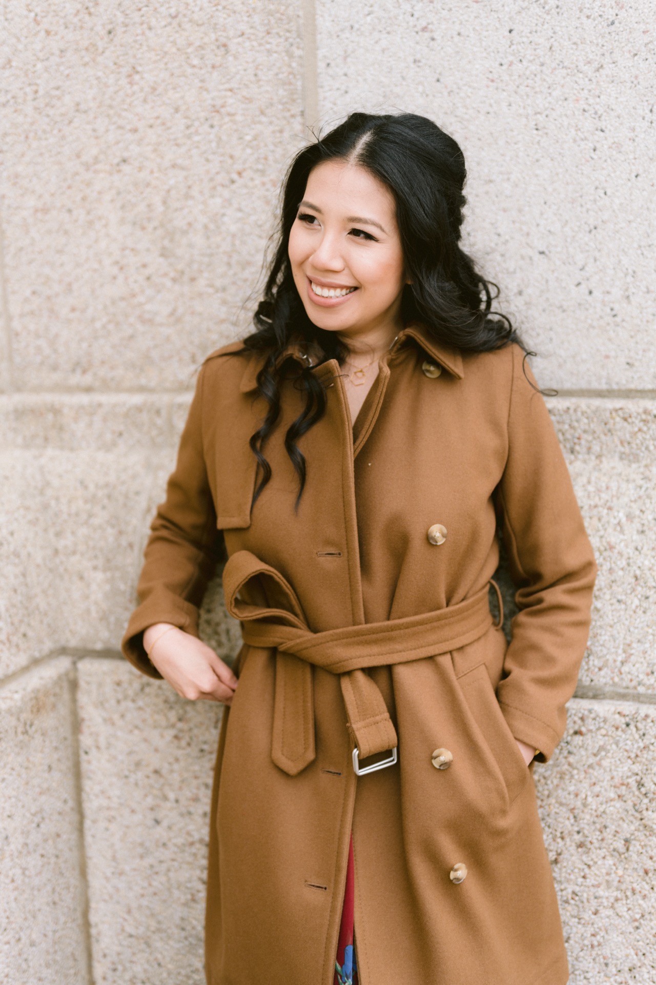 A smiling woman wearing a brown coat standing against a stone wall.