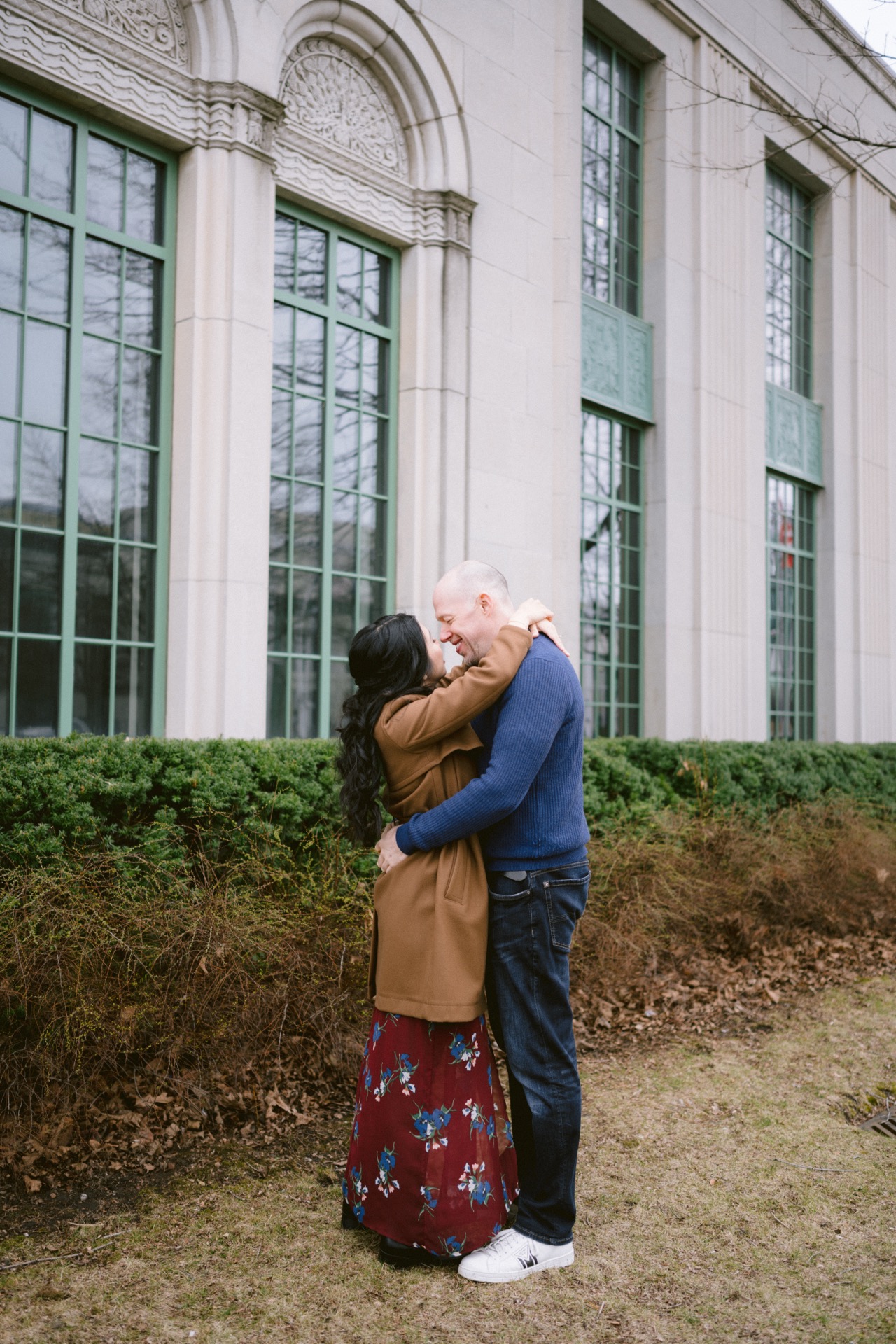 Couple sharing an affectionate embrace outdoors near a building with arched windows.