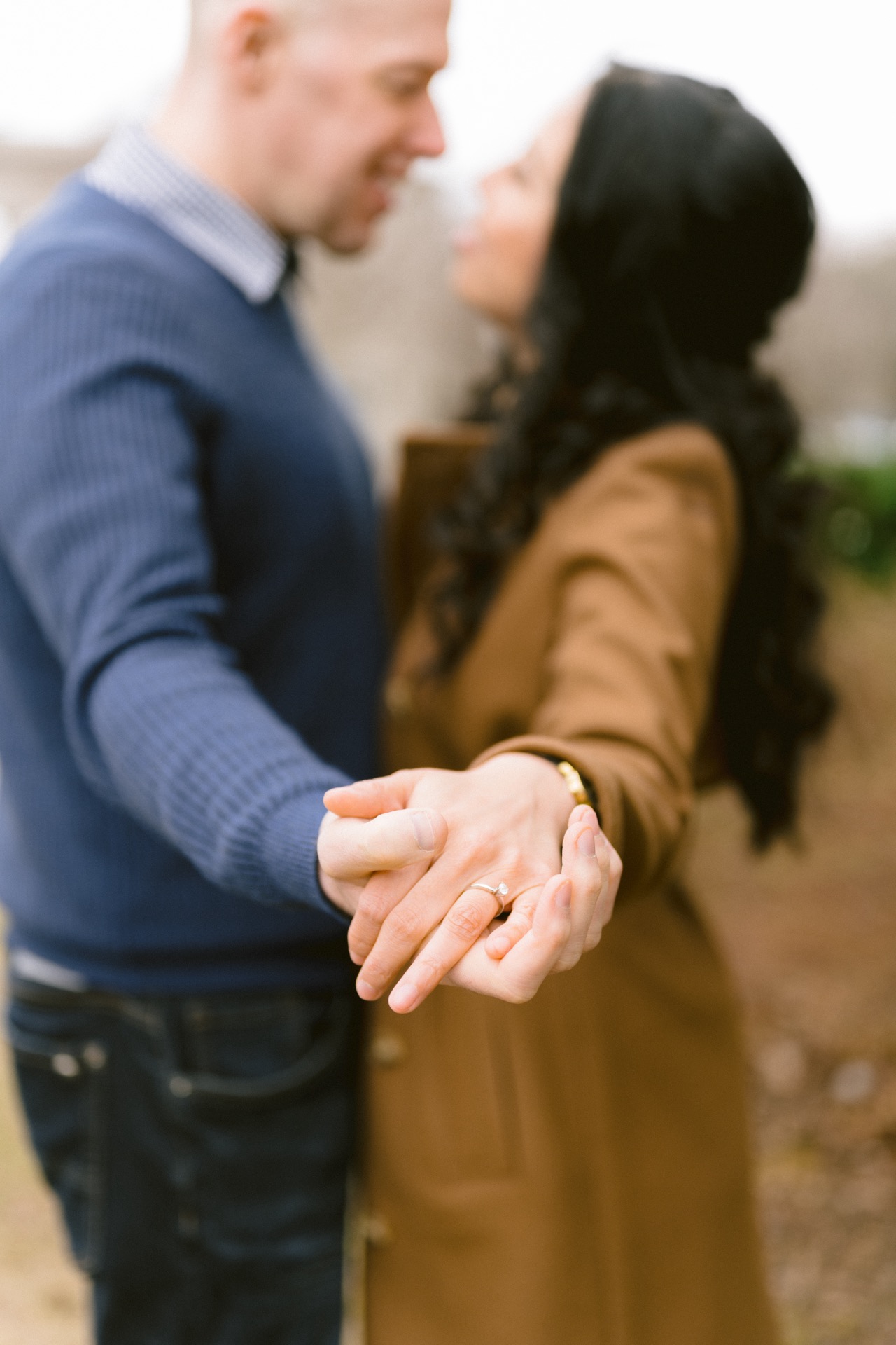A couple showcasing engagement rings while standing close to each other with a blurred background.