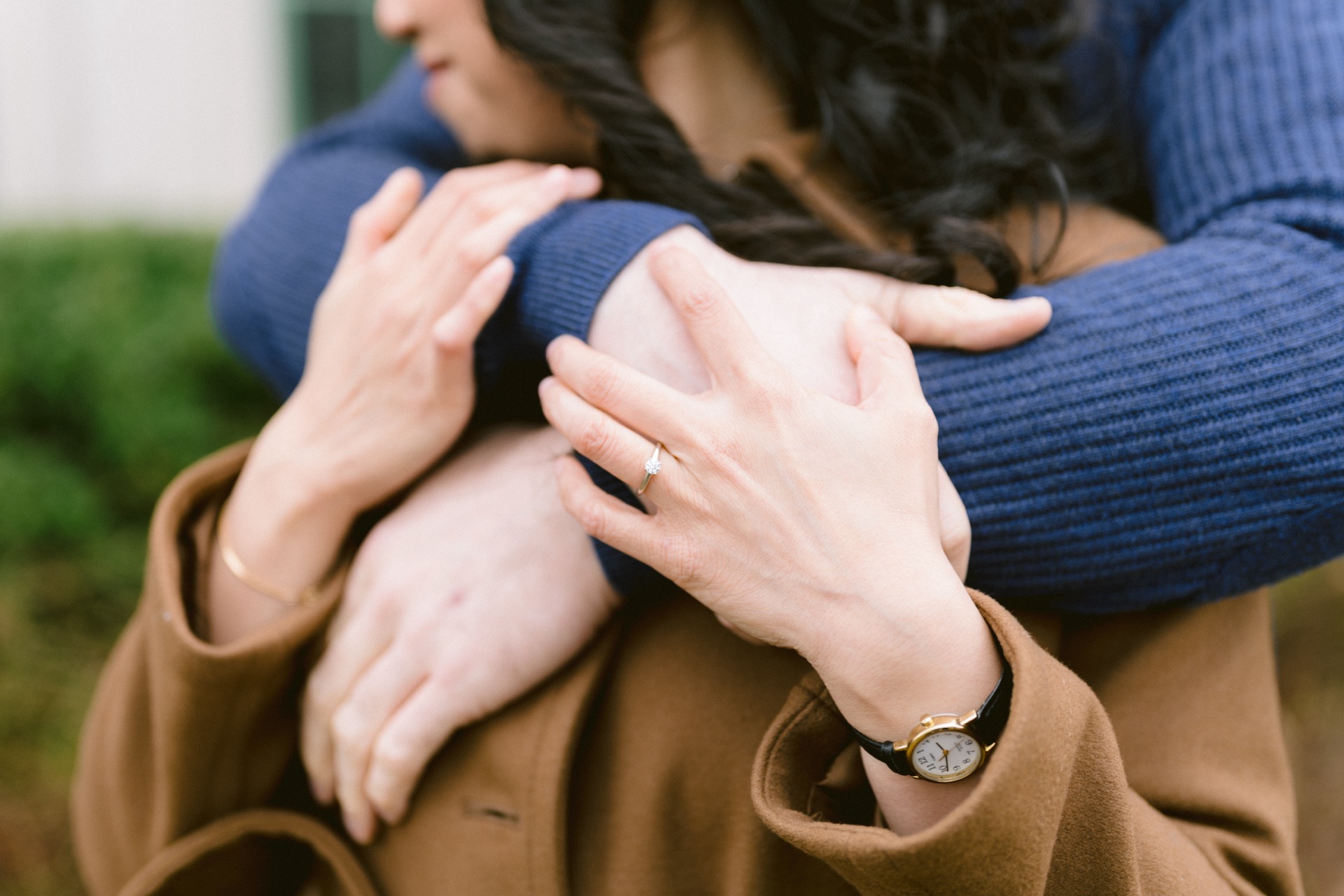 A couple showcasing engagement rings while standing close to each other with a blurred background.