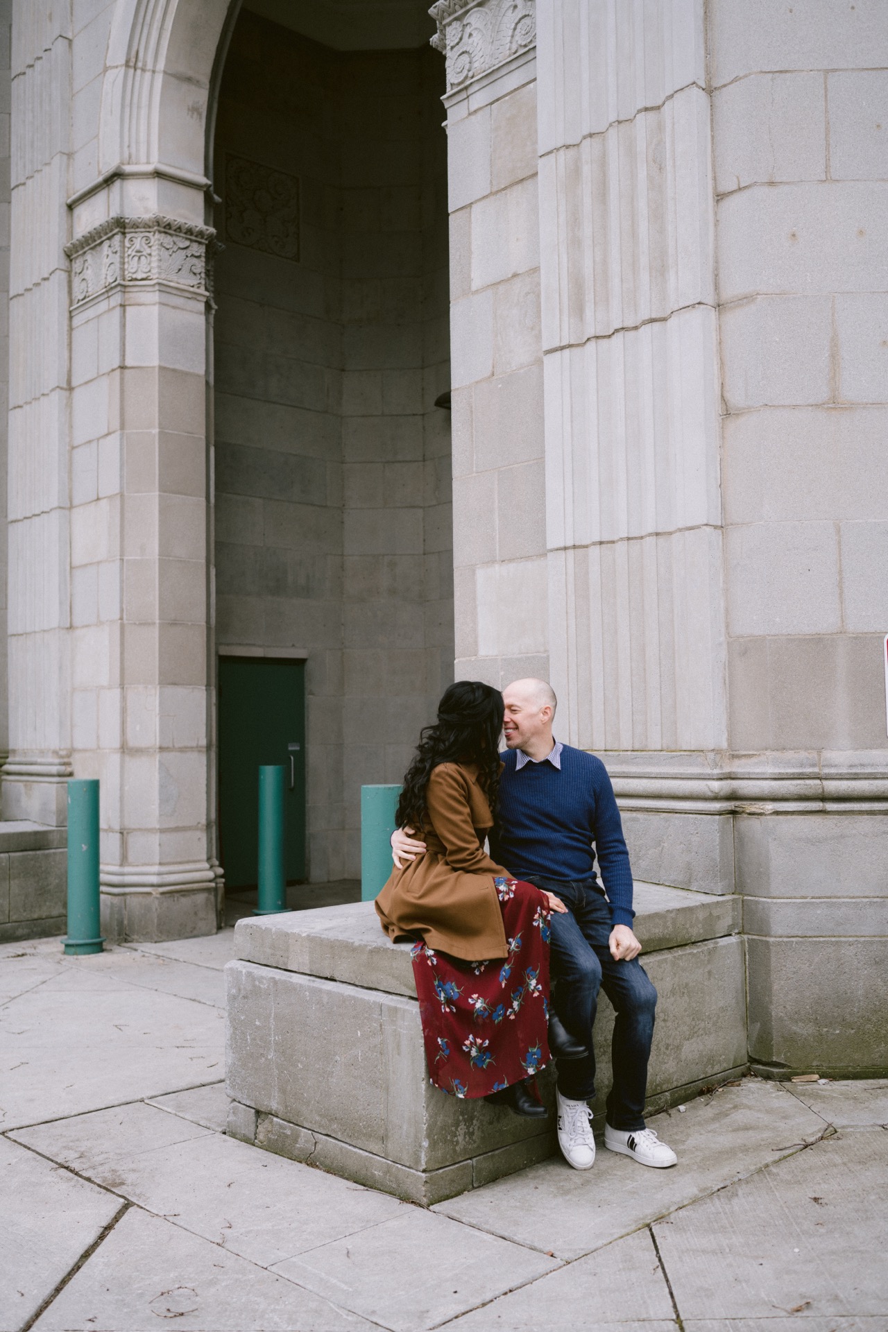 A couple sitting closely together on a stone bench by a building with arches, engaging in a moment of affectionate conversation.