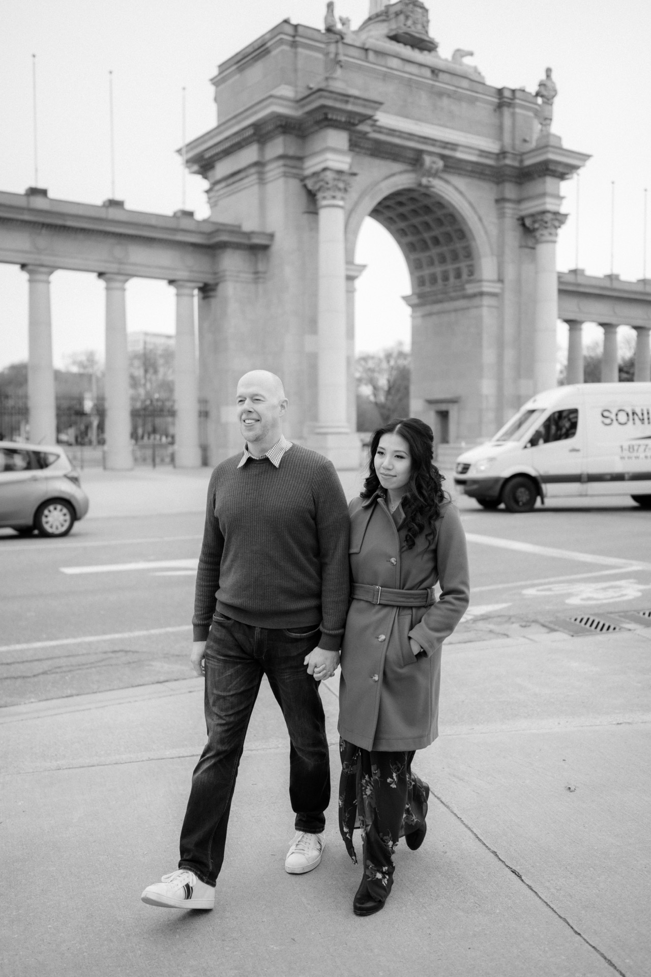 A couple walking hand in hand near an arch monument in a city setting, captured in black and white.