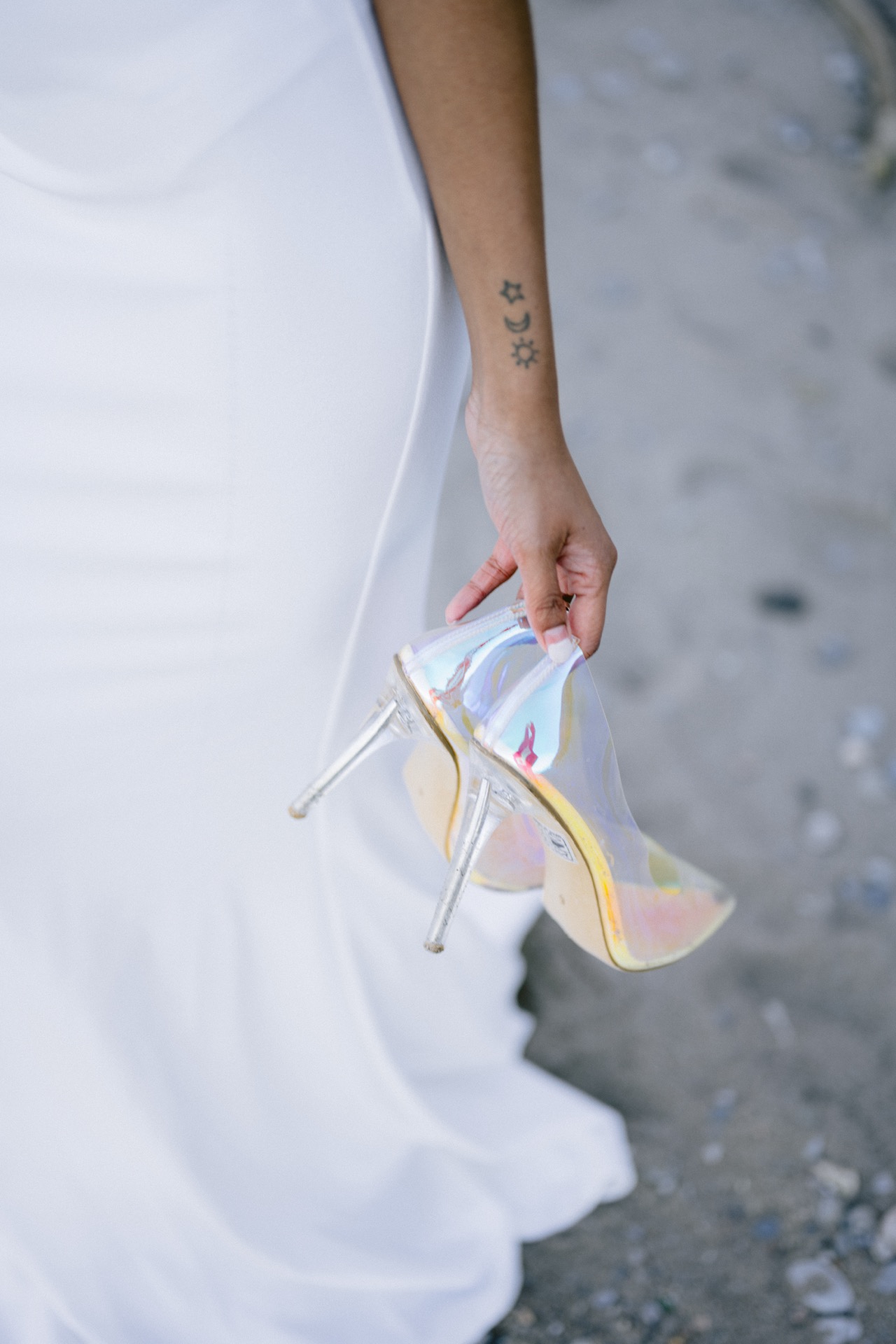 Bride with a tattoo on her hand and carry her high heels while walking on sand.