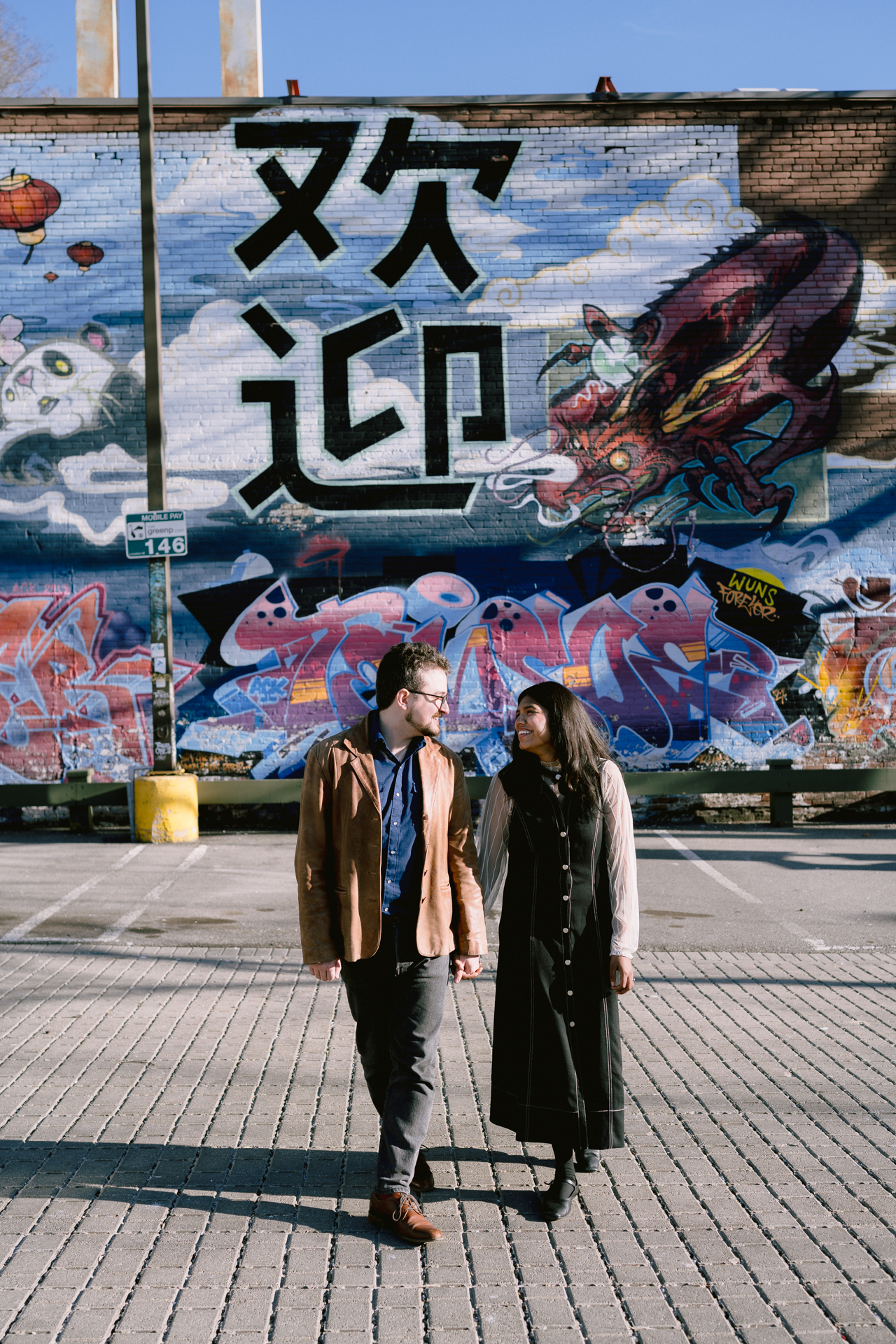 Couple holding hand and walks toward photographer with a huge artistic murals as a background.